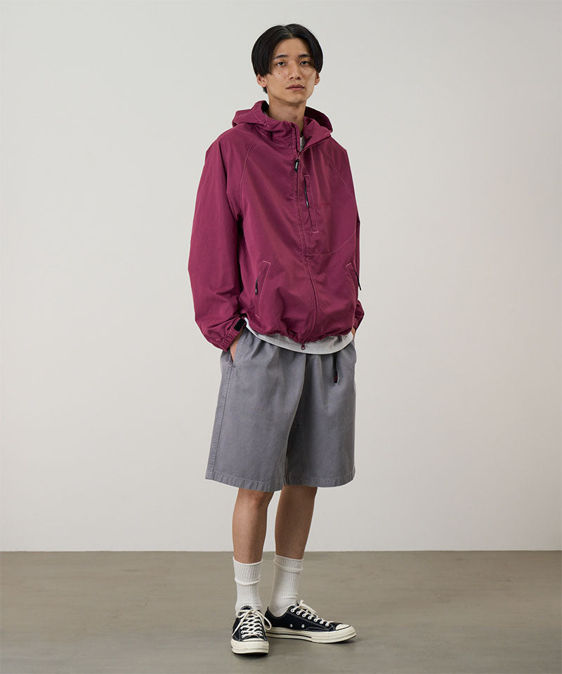Model wearing a burgundy Gramicci Milton Hooded Jacket and grey shorts on a light-tone background.