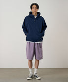 Model wearing a dark navy Gramicci Milton Hooded Jacket and purple shorts on a light-tone background.