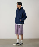 Model wearing a dark navy Gramicci Milton Hooded Jacket and purple shorts on a light-tone background.