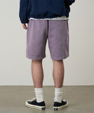 Back view of a model wearing a Gramicci Natural Dyed Jam Short in the color purple on a light-tone background.
