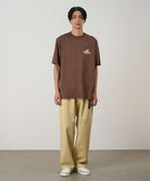Model wearing a Natural Dyed Gramicci Pant Straight Fit in chino and brown shirt on a light-tone background.
