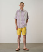Model wearing banana colored Gramicci Nylon Block Shorts and a purple plaid shirt on a light-tone background.