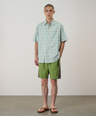 Model wearing jade green Gramicci Nylon Block Shorts and a green plaid shirt on a light-tone background.