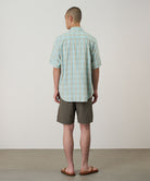 Back view of a model wearing jade green Gramicci Nylon Block Shorts and a green plaid shirt on a light-tone background.