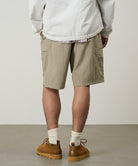 Back view of a model wearing milk tea colored Gramicci Nylon Cliff Short and a white jacket on a light-tone background.