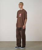 Model wearing walnut Gramicci Milton Pants and a brown shirt on a light-tone background.