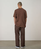 Back view of a model wearing walnut Gramicci Milton Pants and a brown shirt on a light-tone background.