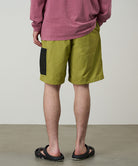 Back view of a model wearing field green Gramicci Guide Shorts and a red shirt on a light-tone background.