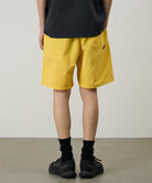 Back view of a model wearing banana colored Gramicci Nylon Reef Shorts and a black shirt on a light-tone background.