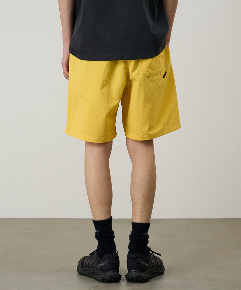 Back view of a model wearing banana colored Gramicci Nylon Reef Shorts and a black shirt on a light-tone background.