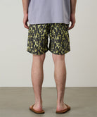 Back view of a model wearing olive camo Gramicci Nylon Reef Shorts and a lavender shirt on a light-tone background.