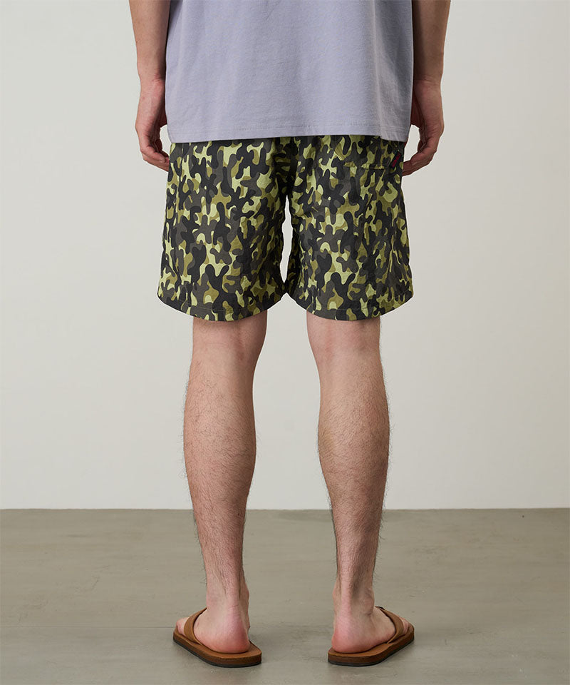 Back view of a model wearing olive camo Gramicci Nylon Reef Shorts and a lavender shirt on a light-tone background.