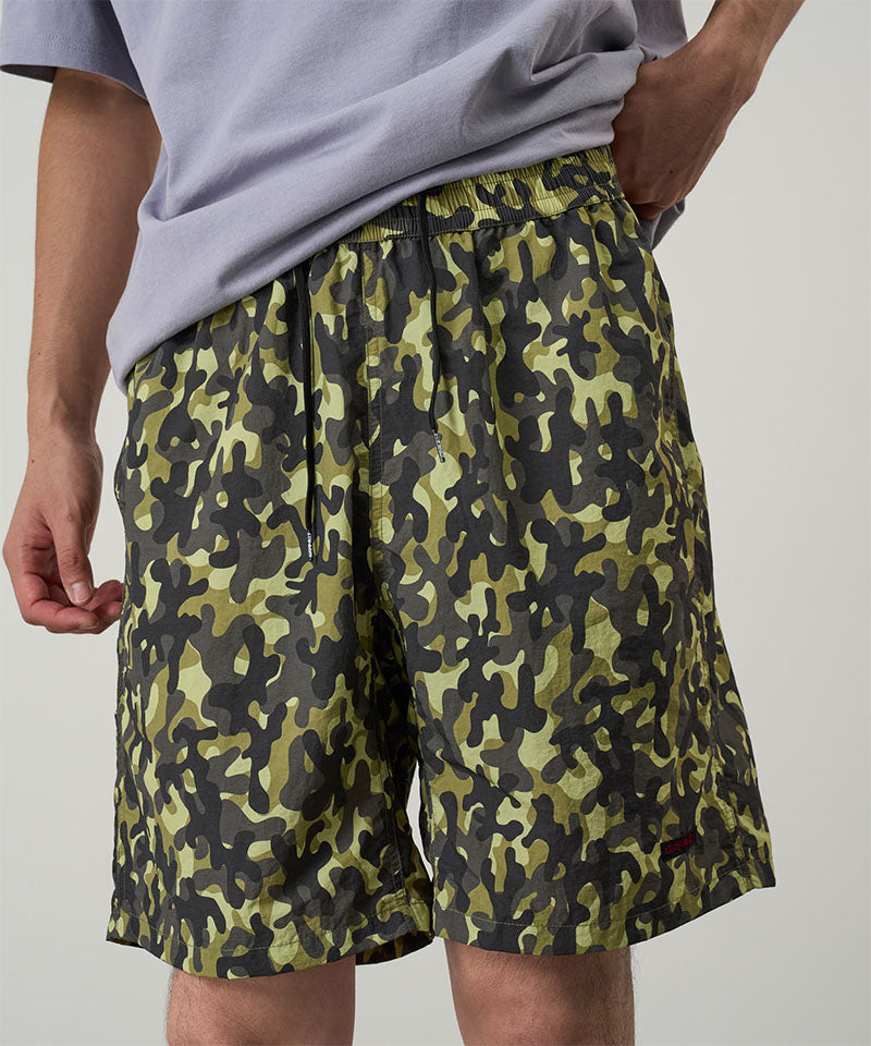 Model wearing olive camo Gramicci Nylon Reef Shorts and a lavender shirt on a light-tone background.