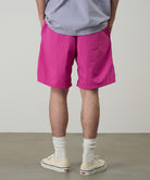 Back view of a model wearing bright plum colored Gramicci Nylon Packable G-Short and a lavender shirt on a light-tone background.