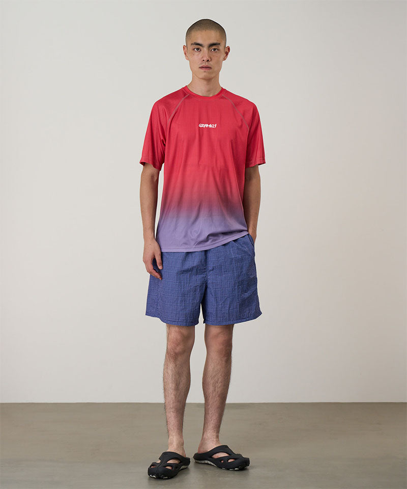 Model wearing a red gradient Gramicci UPF Shield S/S Top with a white Gramicci graphic and blue shorts on a light-tone background.
