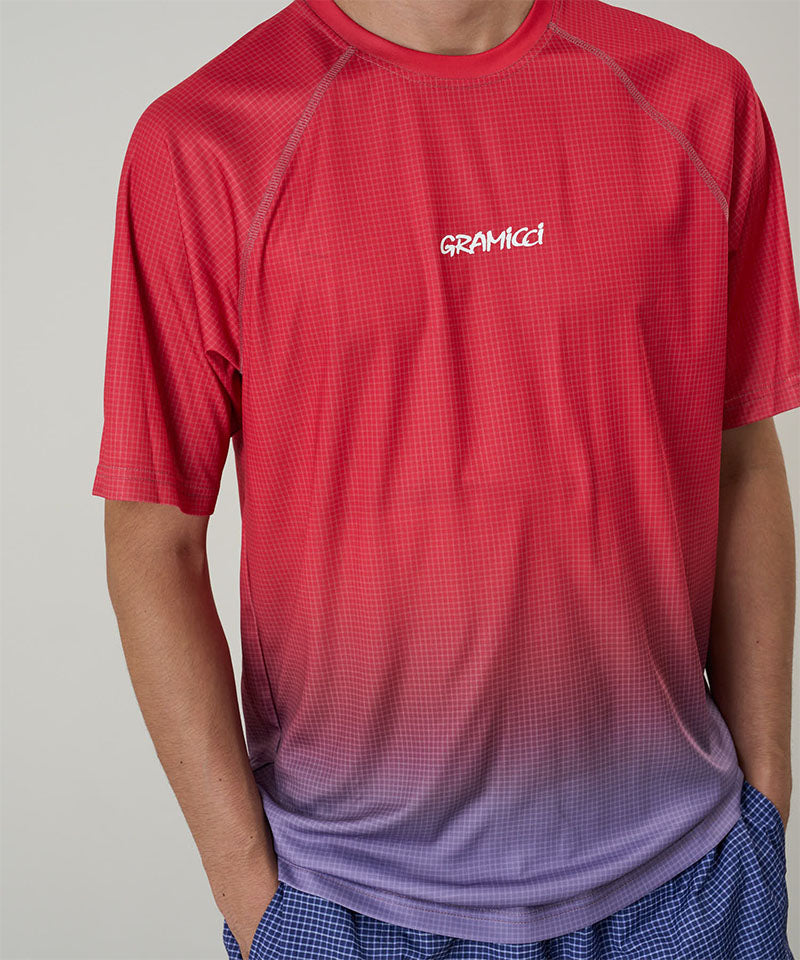 Model wearing a Gramicci red gradient UPF Shield S/S Top with a white Gramicci graphic against a light-tone background.
