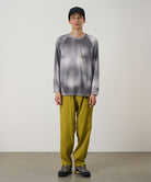Model wearing a black tie-dye Gramicci L/S 1st Layer shirt with grey graphic and yellow pants on a light-tone background.