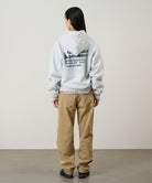 Back view of a model wearing a heather ash Gramicci G-Sail Zip Hooded Sweatshirt with a teal graphic and khaki pants on a light-tone background.