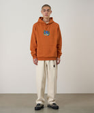 Model wearing the Gramicci Blueberry Hooded Sweatshirt with blueberry graphic in orange and cream colored pants on a light tone background.
