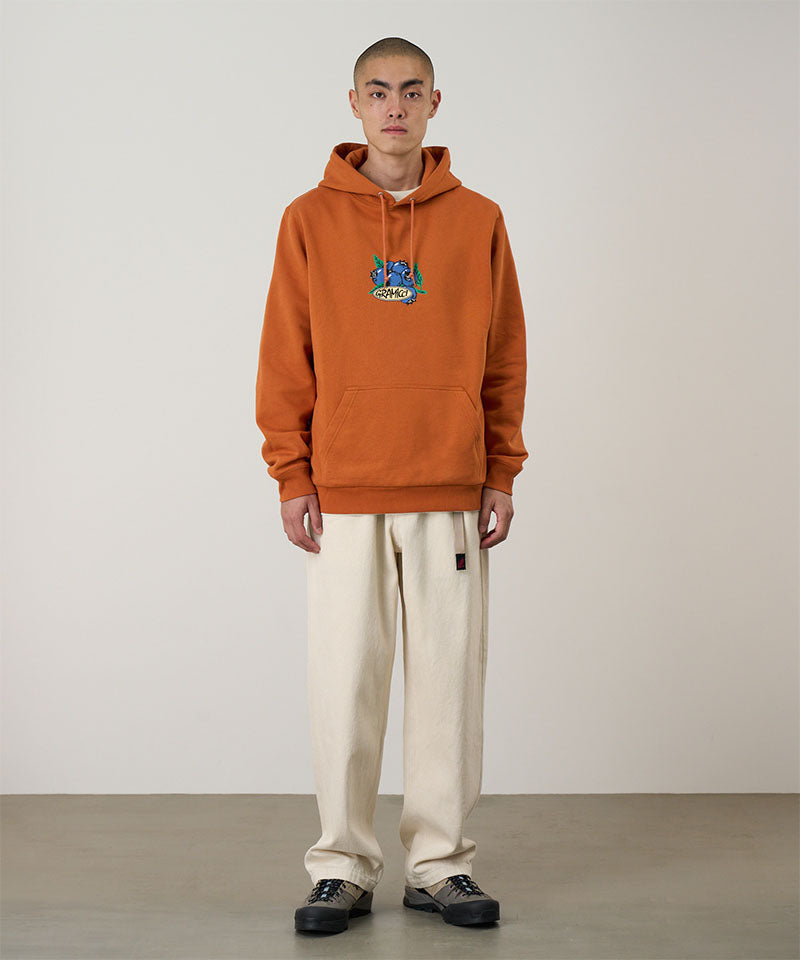 Model wearing the Gramicci Blueberry Hooded Sweatshirt with blueberry graphic in orange and cream colored pants on a light tone background.
