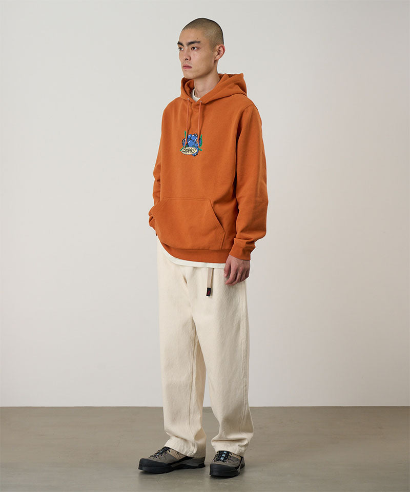 Model wearing the Gramicci Blueberry Hooded Sweatshirt with blueberry graphic in orange and cream colored pants on a light tone background.
