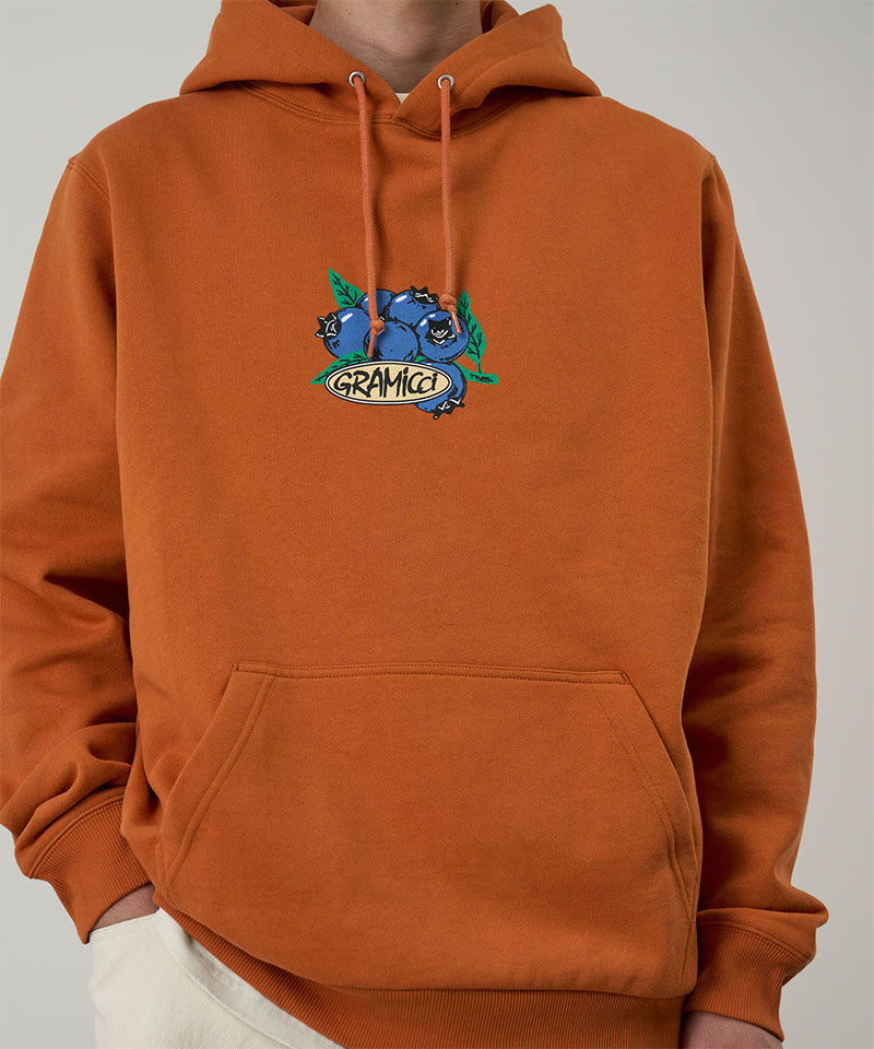 Gramicci Blueberry Hooded Sweatshirt with blueberry graphic in orange worn by a model against a light tone background.
