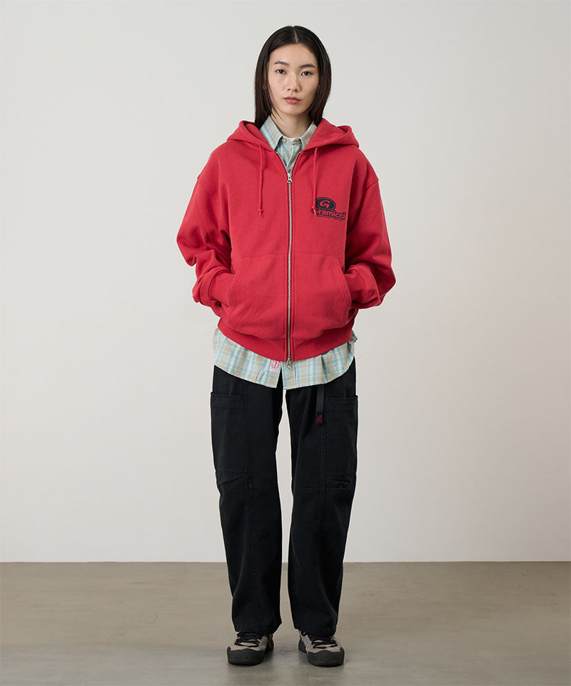 Model wearing a crimson red Gramicci '82 Zip Hooded Sweatshirt with a black graphic and green pants on a light tone background.

