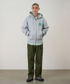 Model wearing a heather grey Gramicci '82 Zip Hooded Sweatshirt with a green graphic and green pants on a light tone background.
