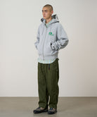 Model wearing a heather grey Gramicci '82 Zip Hooded Sweatshirt with a green graphic and green pants on a light tone background.
