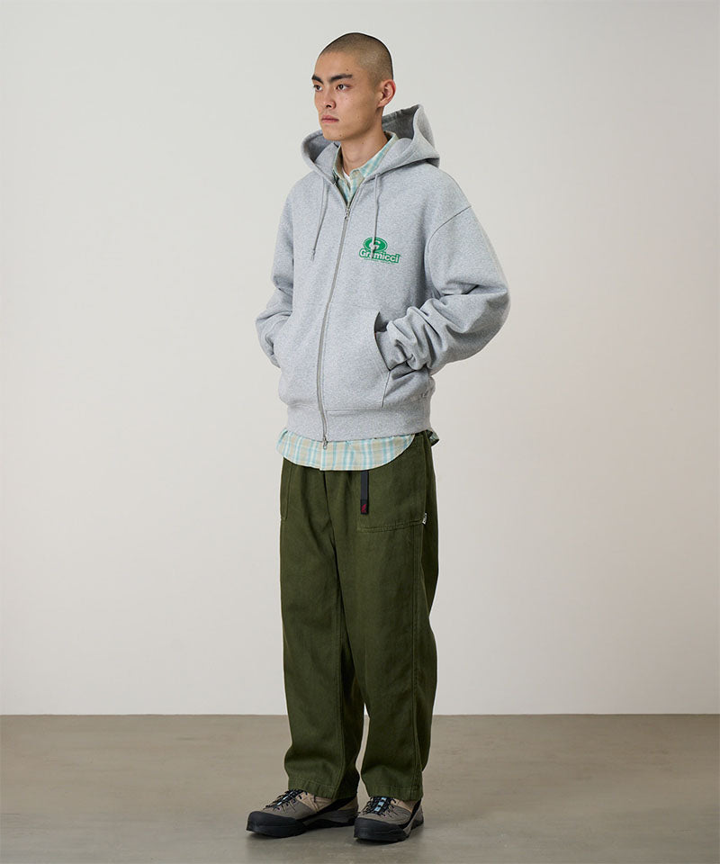 Model wearing a heather grey Gramicci '82 Zip Hooded Sweatshirt with a green graphic and green pants on a light tone background.

