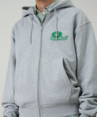 Model wearing a Gramicci heather grey '82 Zip Hooded Sweatshirt with green graphic against a light tone background.
