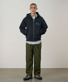 Model wearing a vintage black Gramicci '82 Zip Hooded Sweatshirt with a blue graphic and green pants on a light tone background.
