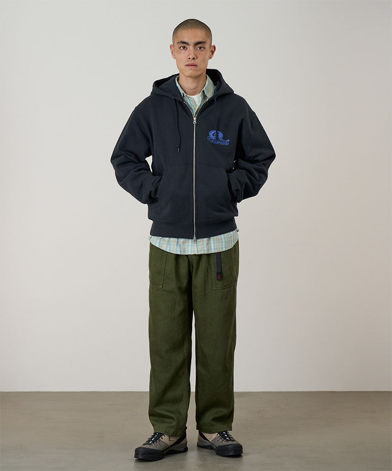 Model wearing a vintage black Gramicci '82 Zip Hooded Sweatshirt with a blue graphic and green pants on a light tone background.
