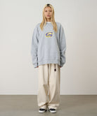 Model wearing a heather grey Gramicci G-Scribble Sweatshirt with a purple and yellow graphic and beige pants on a light tone background.