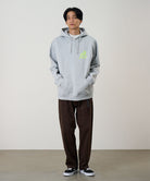Model wearing a heather grey Gramicci Running Man Hooded Sweatshirt with a bright lime running man graphic and olive shorts on a light tone background.