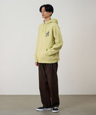 Model wearing a lime Gramicci Running Man Hooded Sweatshirt with a grey running man graphic and brown pants on a light tone background.