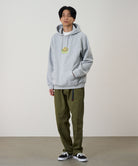Model wearing a heather grey Gramicci Turtle Hooded Sweatshirt with a turtle graphic and olive pants on a light tone background.