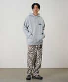 Model wearing a heather grey Gramicci Half Dome Hooded Sweatshirt with a beige and green graphic and brown patterned pants on a light tone background.