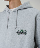 Model wearing a Gramicci heather grey Half Dome Hooded Sweatshirt with beige and green graphic against a light tone background.