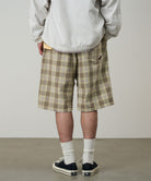 Back view of a model wearing shadow plaid colored Gramicci Swell Short Shadow Plaid and a off-white jacket on a light-tone background.