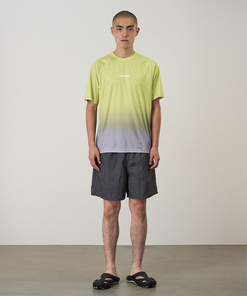 Model wearing black checked Gramicci Grid Swim Shorts and a lime and grey gradient t-shirt on a light-tone background.

