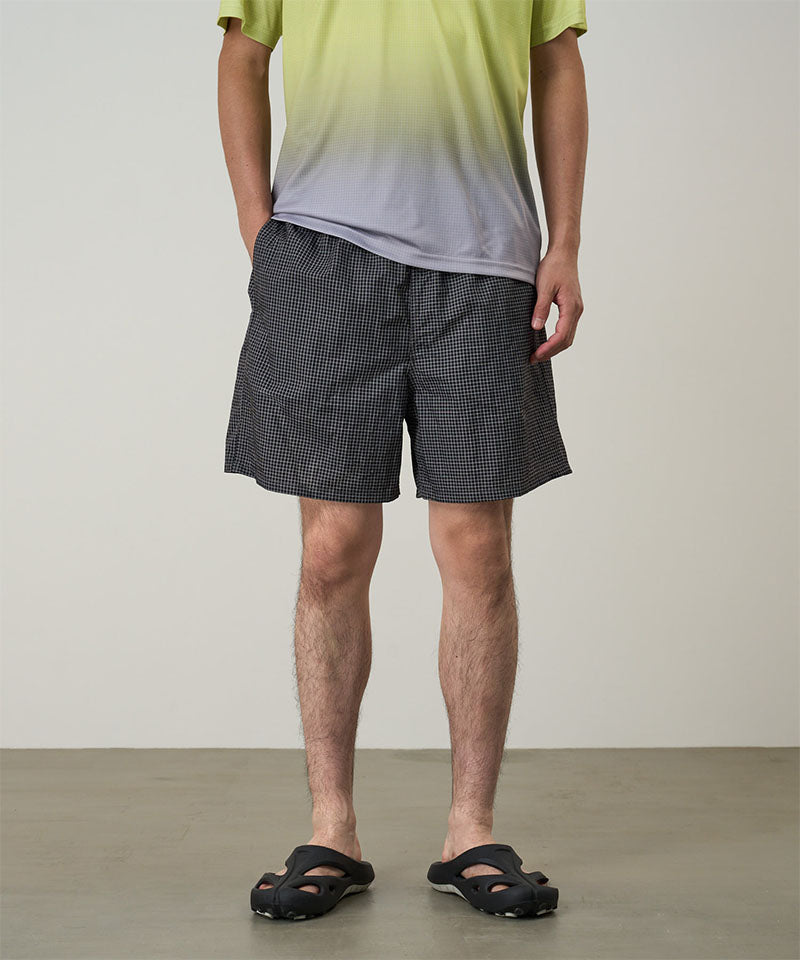 Model wearing black checked Gramicci Grid Swim Shorts and a lime and grey gradient t-shirt on a light-tone background.

