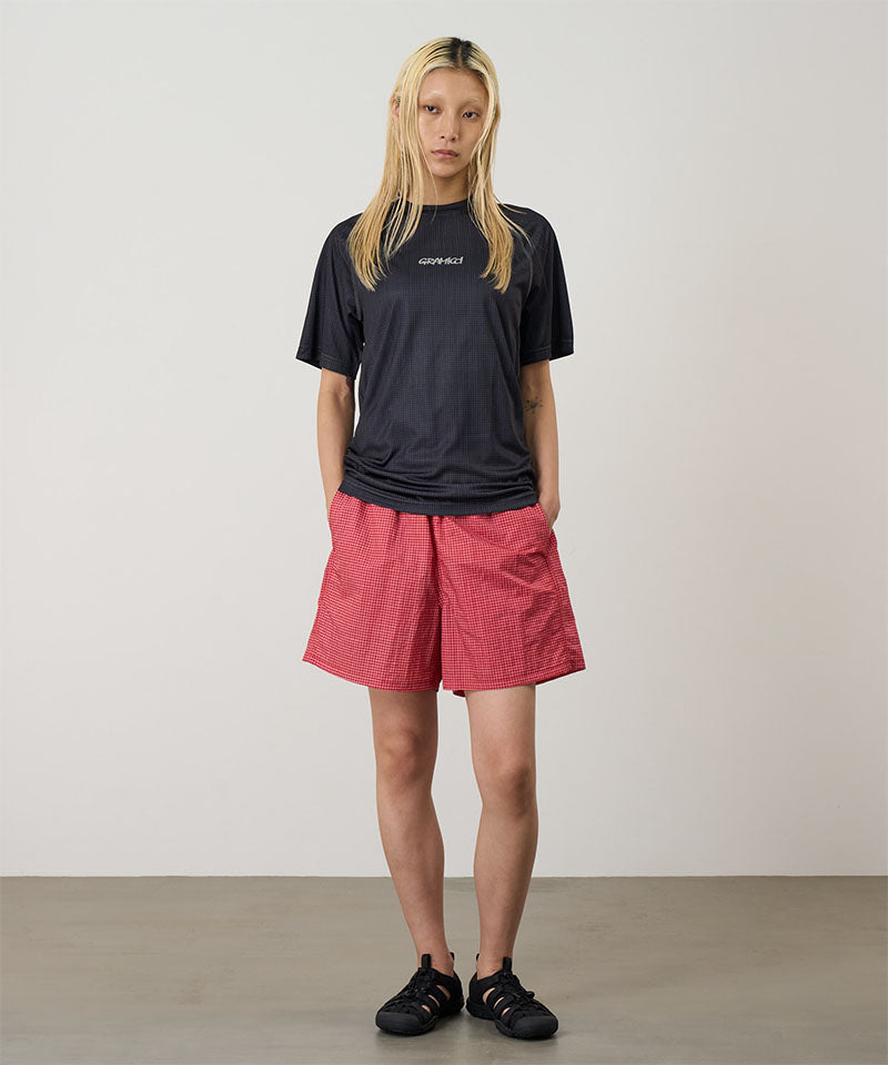 Model wearing red checked Gramicci Grid Swim Shorts and a black t-shirt on a light-tone background.
