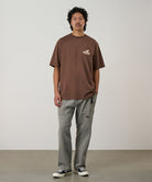 Model wearing a brown Gramicci Greenicci Tee with a  graphic and grey shorts on a light tone background.
