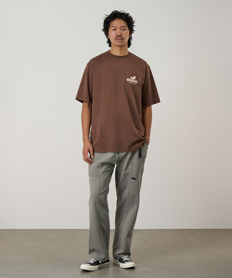 Model wearing a brown Gramicci Greenicci Tee with a  graphic and grey shorts on a light tone background.
