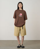 Model wearing a brown Gramicci Pomegranate Tee with a a graphic and beige shorts on a light tone background.
