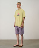Model wearing a lime green Gramicci Pomegranate Tee with a a graphic and purple shorts on a light tone background.
