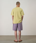 Back view of a model wearing a lime green Gramicci Pomegranate Tee and purple shorts on a light tone background.
