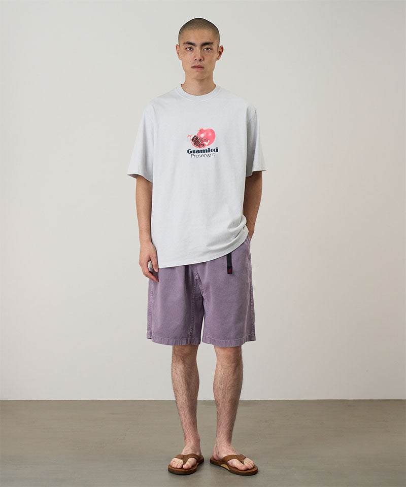 Model wearing a smoke colored Gramicci Pomegranate Tee with a a graphic and purple shorts on a light tone background.
