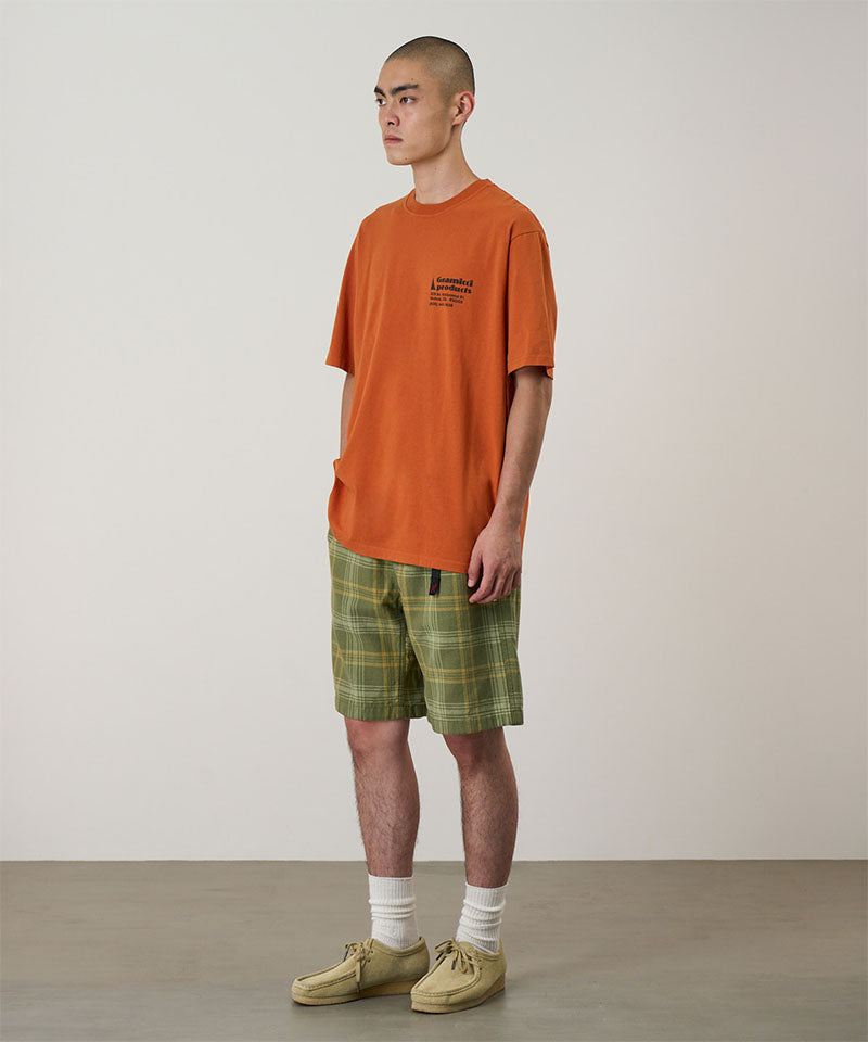 Model wearing a orange Gramicci G-Sail Tee with black graphic and green plaid pants on a light-tone background.